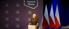 Mężczyzna w mundurze stojąc przy mównicy przemawia za nim ustawione są trzy flagi Polski oraz baner z napisem Konwent Morski oraz logo.