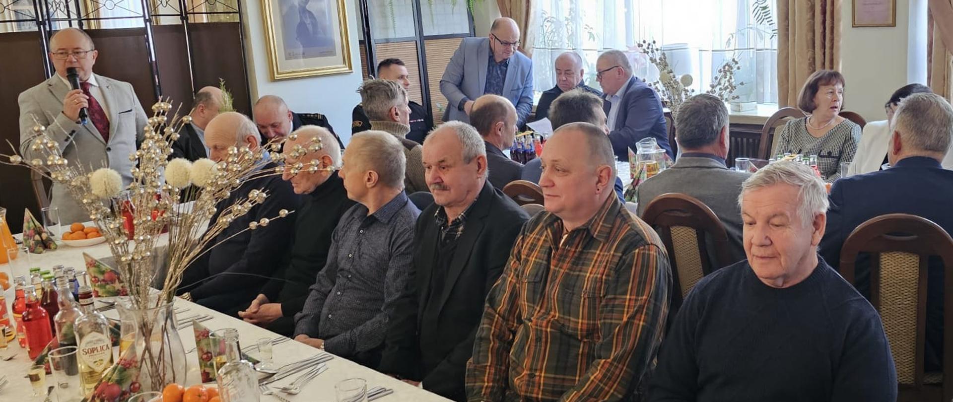 Uczestnicy spotkania okolicznościowego siedzący przy długich stołach w sali, w tym funkcjonariusze Państwowej Straży Pożarnej oraz zaproszeni goście