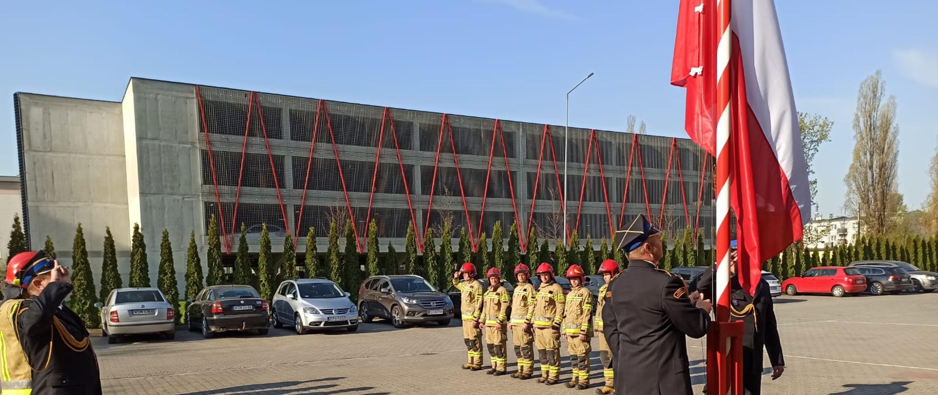 Flaga Rzeczpospolitej Polskiej jest wciągana na maszt strażacy oddają salutują.