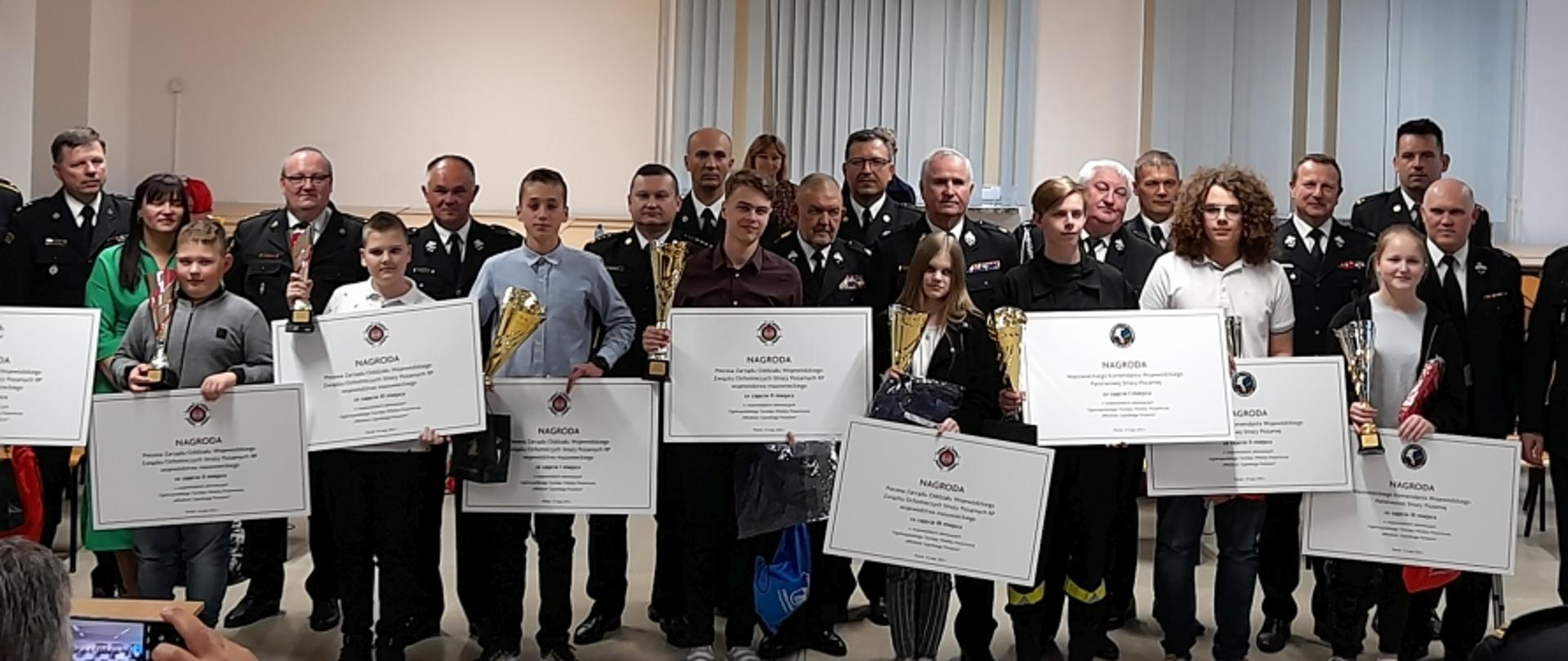 Zdjęcie grupowe uczestników konkursu, trzymających dyplomy, puchary i nagrody rzeczowe. Za nimi stoją strażacy PSP i OSP w mundurach wyjściowych.