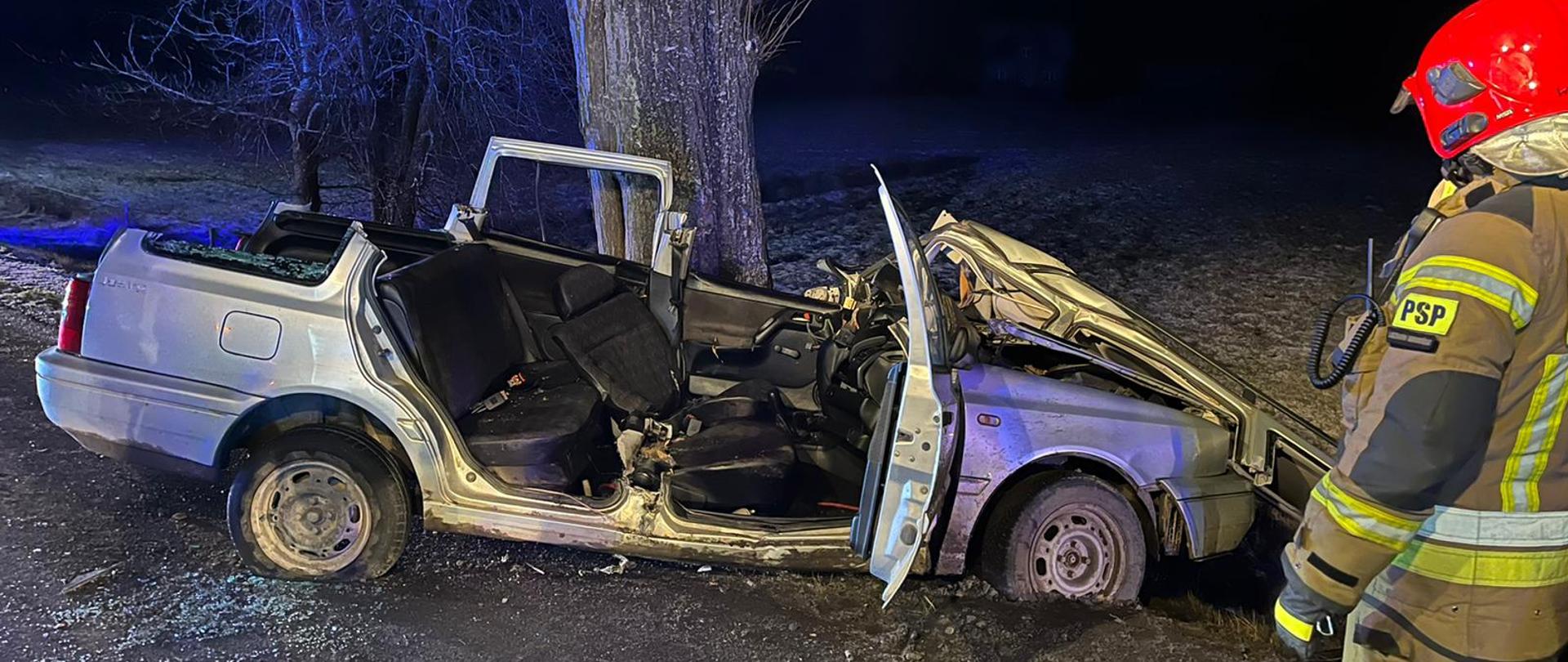 Zdjęcie przedstawia wrak pojazdu po czołowym uderzeniu z przydrożnym drzewem oraz strażaka PSP