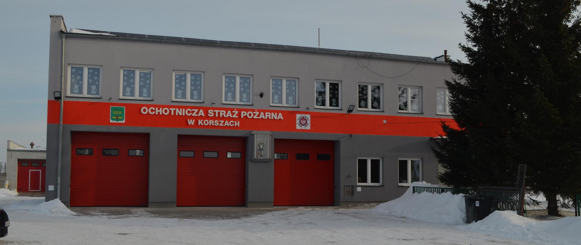 Zdjęcie przedstawia budynek OSP Korsze po termomodernizacji. Zdjęcie zrobione od strony czerwonych bram garażowych z nową elewacją.
