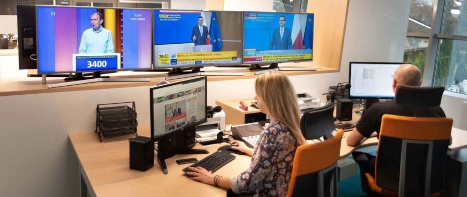 Młoda kobieta i młody mężczyzna siedzą przed komputerami. Przed nimi, trzy ekrany telewizorów z włączonymi rożnymi programami informacyjnymi.