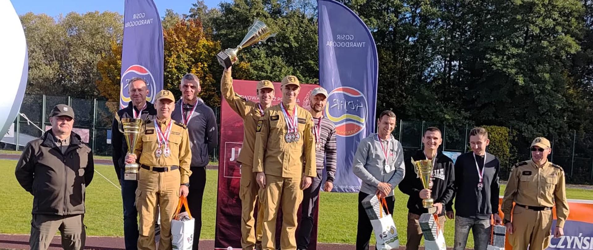Uczestnicy zawodów sportowych pozują do wspólnego zdjęcia podczas ceremonii wręczenia nagród. Zawodnicy w umundurowaniu stoją na podium, unosząc trofea, a pozostali uczestnicy prezentują zdobyte nagrody. W tle widoczne są banery promocyjne, drzewa oraz bieżnia sportowa.