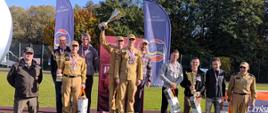 Uczestnicy zawodów sportowych pozują do wspólnego zdjęcia podczas ceremonii wręczenia nagród. Zawodnicy w umundurowaniu stoją na podium, unosząc trofea, a pozostali uczestnicy prezentują zdobyte nagrody. W tle widoczne są banery promocyjne, drzewa oraz bieżnia sportowa.