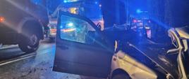 Zdjęcie przedstawia wrak pojazdu po czołowym uderzeniu z przydrożnym drzewem oraz służby 