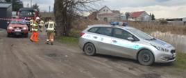 Na drodze gruntowej obok stawu stoi radiowóz policyjny, a za nim auto terenowe i wóz ratowniczo-gaśniczy z Komendy Powiatowej PSP w Rawiczu. Obok pojazdów pożarniczych przechodzą strażacy. Mają na sobie ubrania specjalne w kolorze piaskowym. Dwóch z nich założone ma również pomarańczowe wodery. Staw jest porośnięty trzciną. Z lewej strony widoczne sa zabudowania.