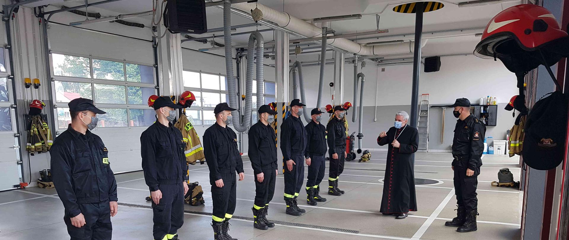 Zdjęcie zrobione w dzień w garażu JRG Sandomierz. Na zdjęciu widać siedmiu strażaków, którzy stoją w szeregu oraz księdza biskupa i komendanta powiatowego.
W dalszej części zdjęcia widać ubrania i hełmy strażackie.
