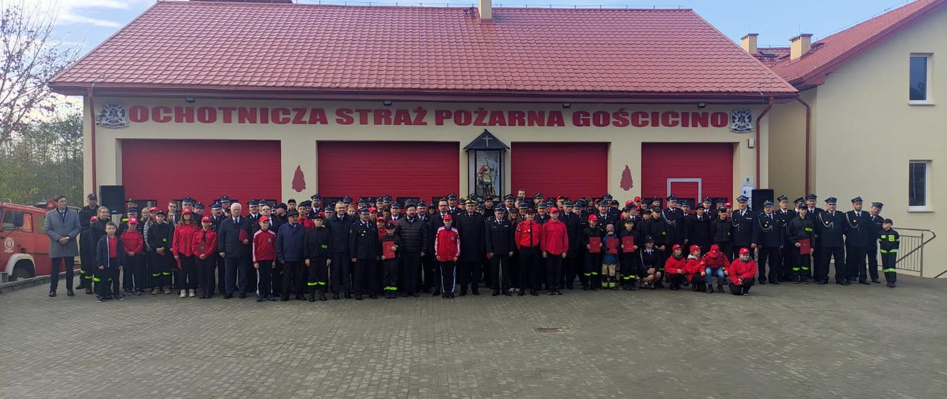 Strażacy Państwowej Straży Pożarnej przedstawiciele ochotniczych straży pożarnych oraz młodzieżowych drużyn pożarniczych stoją obok siebie przed bramami wyjazdowymi.