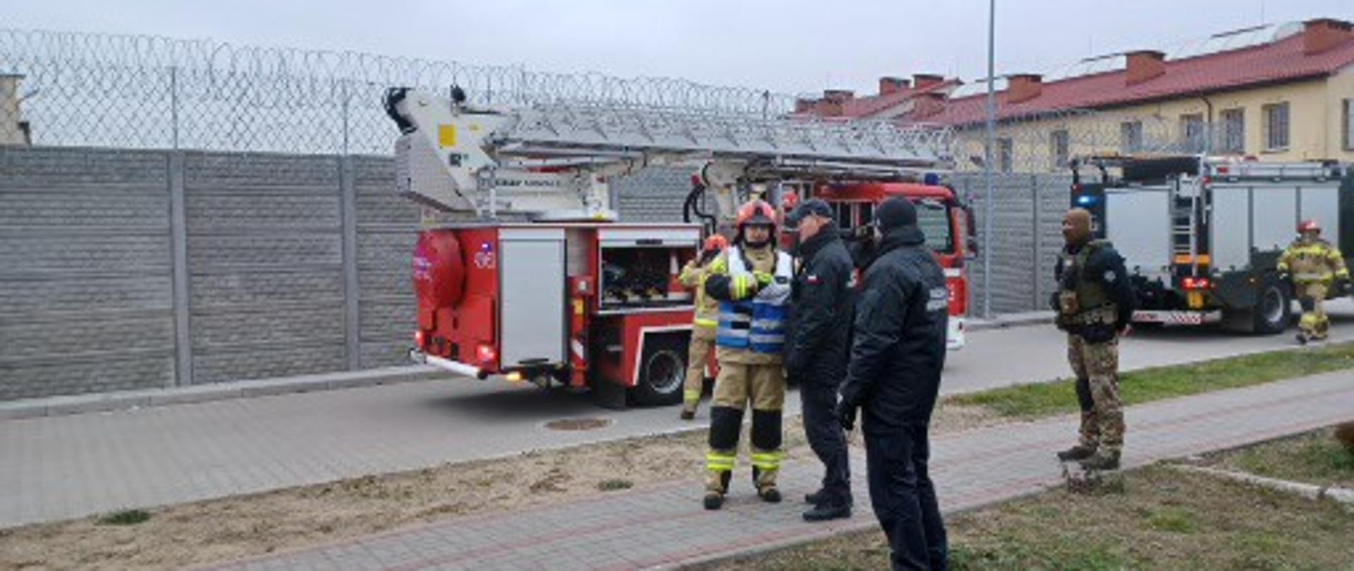 Na pierwszym planie widać kierującego działaniem ratowniczym zbierającego informacje od dowódcy zmiany w zakładzie karnym. Widać również innych strażaków i funkcjonariusz służby więziennej. W tle stoją samochody pożarnicze, widać również fragment ogrodzenia z drutem kolczastym oraz budynek. 