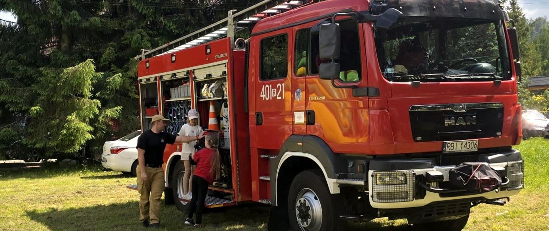 Zdjęcie przedstawia samochód gaśniczy przy którym stoi strażak i dwie dziewczynki oglądają sprzęt który znajduje się na wyposażeniu samochodu. 