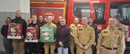Strażacy i pracownicy Komendy Miejskiej PSP w Siemianowicach Śląskich stoją w garażu przed czerwonym wozem strażackim wraz z dyrektorem MOPS. Kilku funkcjonariuszy trzyma duże świąteczne torby z upominkami oznaczone motywami Mikołaja. Wszyscy pozują do wspólnego zdjęcia.