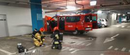 Na zdjęciu widać dwóch funkcjonariuszy PSP w ubraniu specjalnym i hełmie. Osoby znajdują się w garażu podziemnym. Obok ratowników kaseton wężowy. Za ratownikami samochód pozoracji ze straży pożarnej. Posiada sygnalizację ostrzegawczą, jest koloru czerwonego.