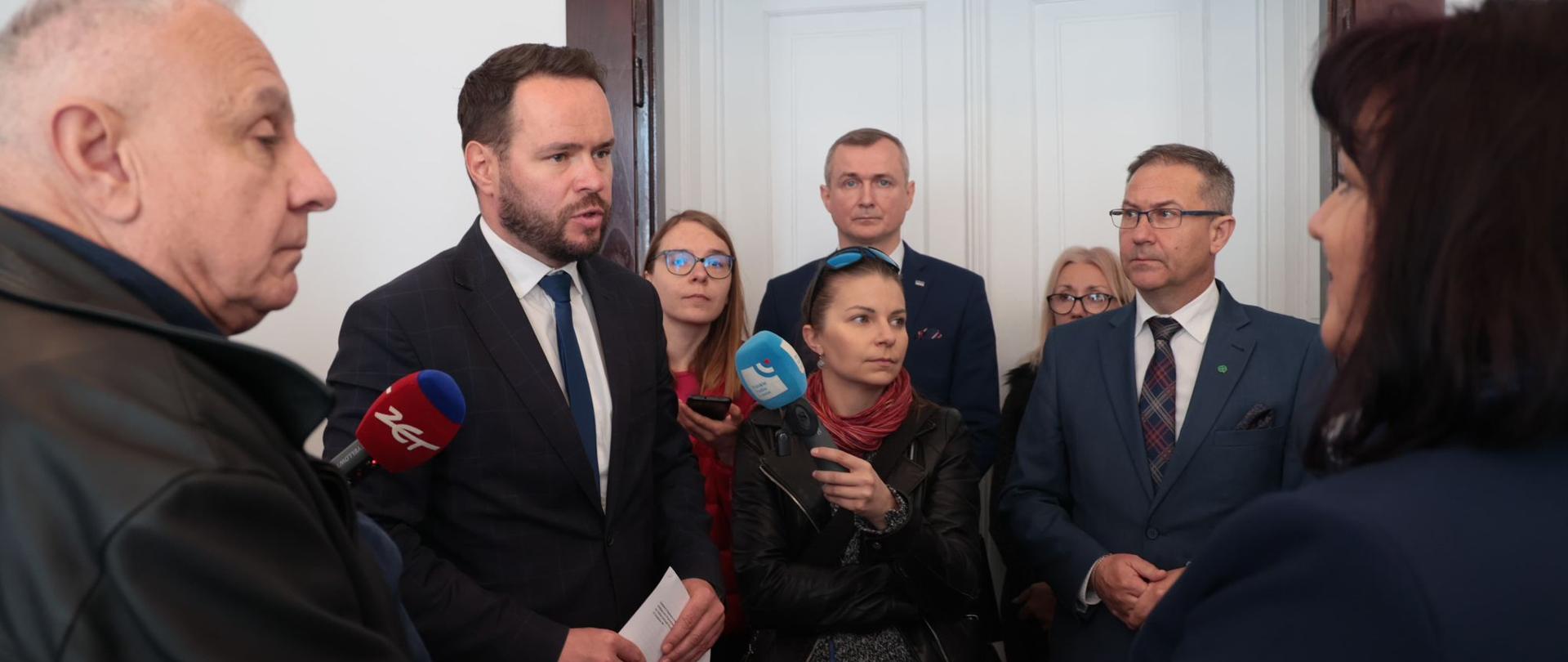 Kilka osób stoi w pomieszczeniu i rozmawia z dziennikarzami trzymającymi mikrofony z logo stacji telewizyjnych. Mężczyzna w ciemnym garniturze i niebieskim krawacie odpowiada na pytania dziennikarzy (to wojewoda lubelski Krzysztof Komorski), trzymając w ręku kartkę papieru. Za nim i obok niego stoją inne osoby, przysłuchując się rozmowie. W tle widać białe drzwi i fragment białej ściany. Oświetlenie jest jasne, a atmosfera wydaje się być oficjalna, jak podczas konferencji prasowej lub wywiadu.