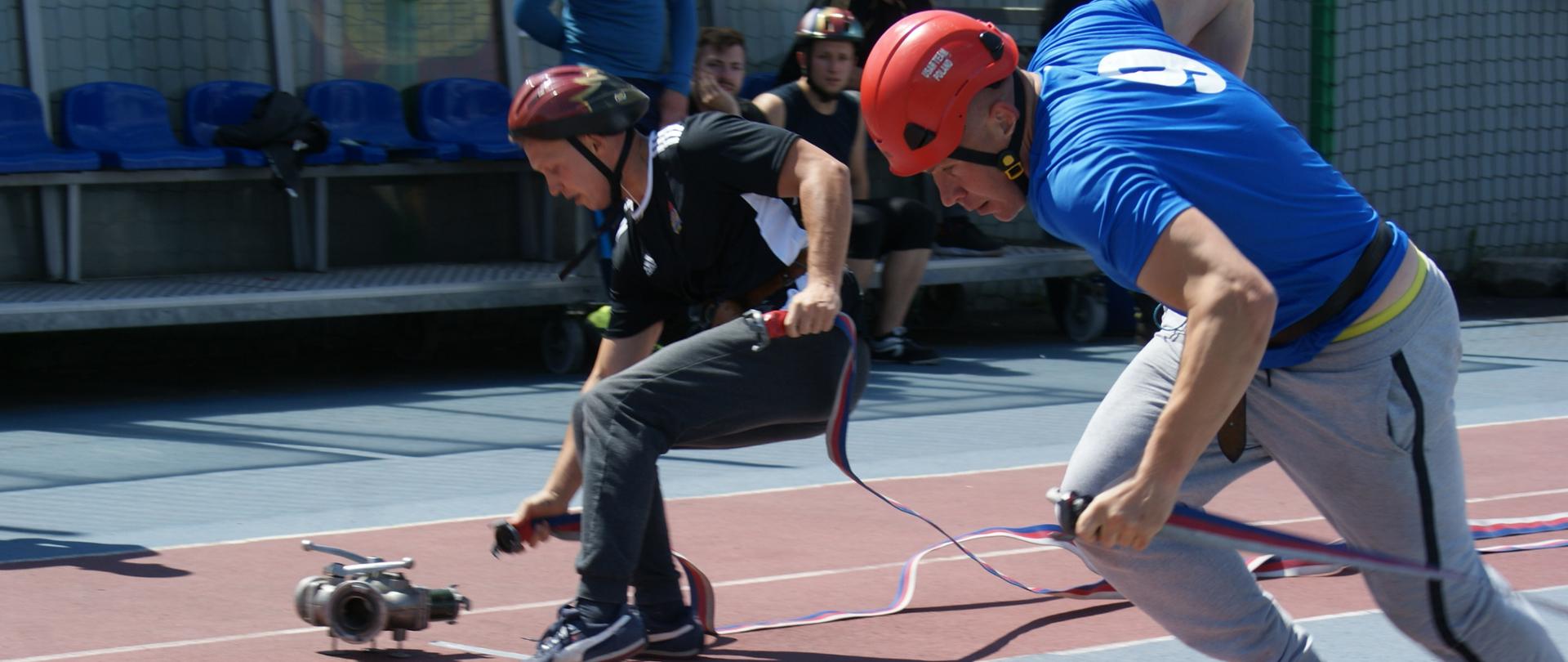 Dwóch strażaków podczas biegu na bieżni lekkoatletycznej w kaskach, jeden podłącza waż strażacki do rozdzielacza