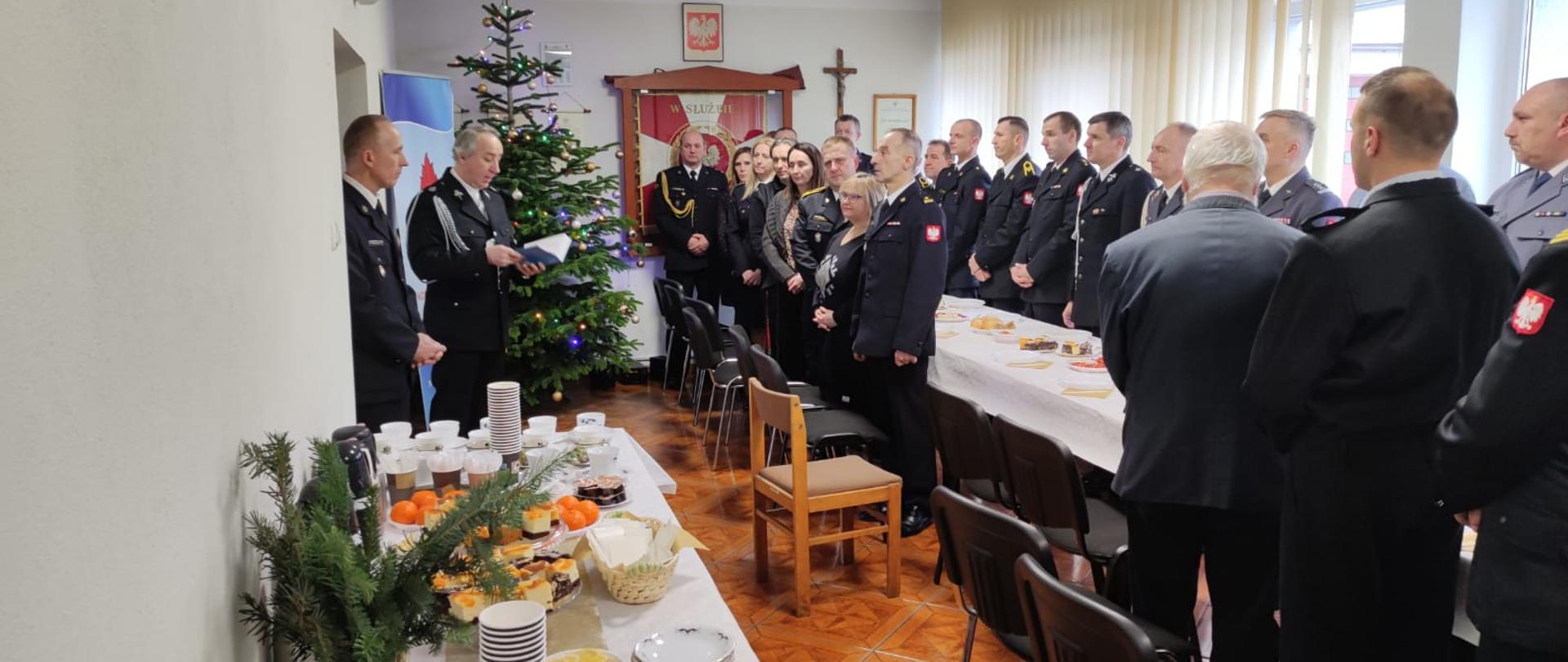 Zdjęcie przedstawia spotkanie wigilijne w KP PSP Świdwin, które odbywa się na świetlicy. Strażacy są ubrani w mundury wyjściowe a ksiądz ich błogosławi.