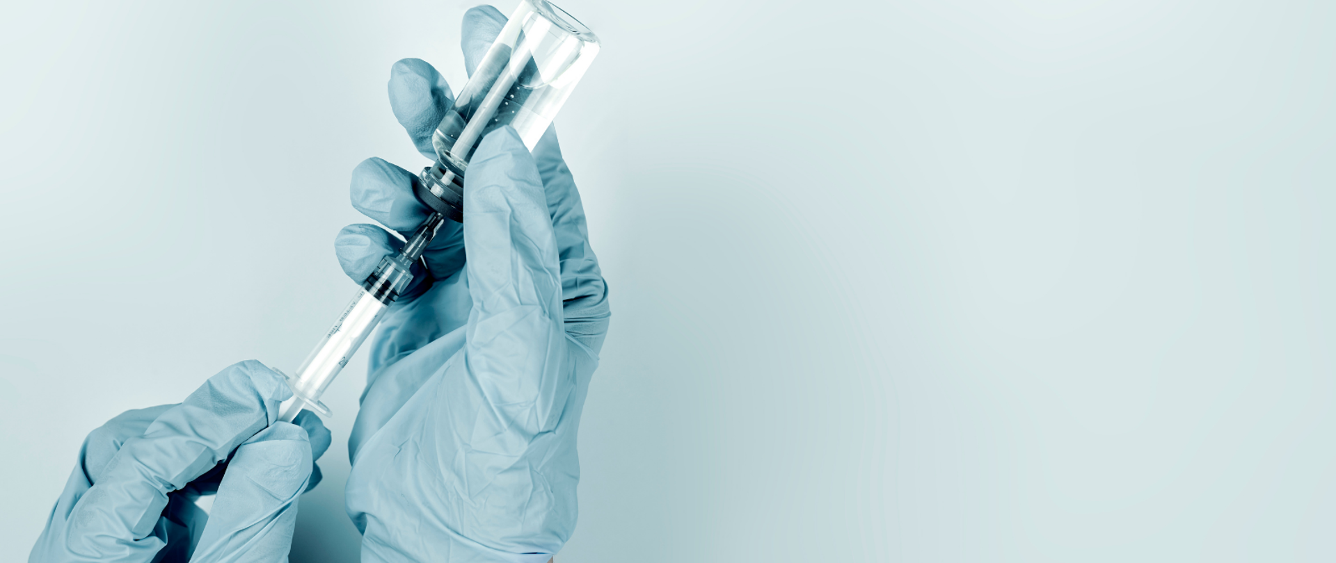 Na bladoniebieskim tle dwie męskie ręce w bladoniebieskich rękawiczkach gumowych trzymają strzykawkę, którą pobierają szczepionkę.