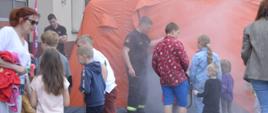 Dzień otwarty w rawickiej komendzie. Przed namiotem pneumatycznym, z kórego wydobywa się dym stoi gromadka dzieci. Jest też strażak, który unosi powłokę namiotu. 