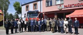 Na zdjęciu osoby kobiety i mężczyźni stojących w szeregu i pozujących do zdjęcia. Kobiety ubrane odświętnie, mężczyźni garnitury. Wśród pozujących widzimy także strażaków, policjantów oraz służbę leśną ubranych w mundury galowe. Za pozującymi widzimy samochód strażacki koloru czerwonego przepasanego biało-czerwoną wstęgą. W tle widzimy także część budynku Jednostki Ratowniczo - Gaśniczej. 