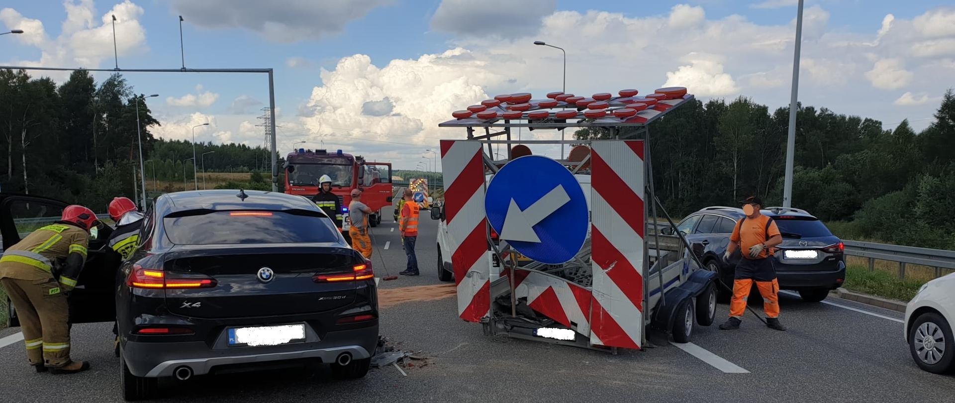 Zdjęcie obrazuje fragment dwupasmowej drogi szybkiego ruchu S7 na której doszło do zderzenia samochodu osobowego marki BMW oraz przyczepki informacyjnej służby drogowej holowanej przez białego busa. Z lewej strony zdjęcia widoczny jest od tyłu czarny samochód marki BMW uczestniczący w zdarzeniu. Przy pojeździe od strony kierowcy dwóch ratowników w ubraniach bojowych i hełmach prowadzą działania ratownicze. Obok samochodu BMW stoi uszkodzona przyczepka informacyjna służb drogowych na której na tle biało czerwonych ukośnych pasów widnieje niebieski znak informacyjny typu kierunek jazdy. Z prawej strony przyczepki widać kolejny samochód osobowy marki Mazda. Za pojazdem BMW widać strażackie pojazdy. Na drodze widoczni są również trzej pracownicy służby drogowej oraz jeden strażak w ubraniu bojowym i hełmie. Na dalszym planie w tle widoczne są drzewa. 