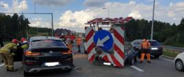Zdjęcie obrazuje fragment dwupasmowej drogi szybkiego ruchu S7 na której doszło do zderzenia samochodu osobowego marki BMW oraz przyczepki informacyjnej służby drogowej holowanej przez białego busa. Z lewej strony zdjęcia widoczny jest od tyłu czarny samochód marki BMW uczestniczący w zdarzeniu. Przy pojeździe od strony kierowcy dwóch ratowników w ubraniach bojowych i hełmach prowadzą działania ratownicze. Obok samochodu BMW stoi uszkodzona przyczepka informacyjna służb drogowych na której na tle biało czerwonych ukośnych pasów widnieje niebieski znak informacyjny typu kierunek jazdy. Z prawej strony przyczepki widać kolejny samochód osobowy marki Mazda. Za pojazdem BMW widać strażackie pojazdy. Na drodze widoczni są również trzej pracownicy służby drogowej oraz jeden strażak w ubraniu bojowym i hełmie. Na dalszym planie w tle widoczne są drzewa. 