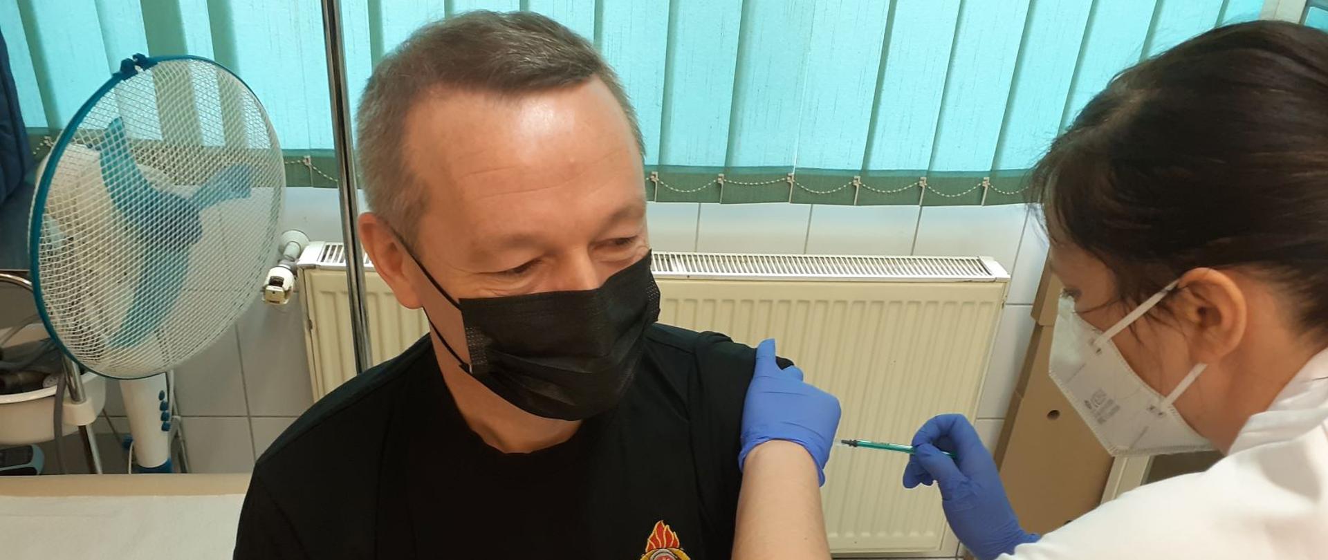 Na zdjęciu widzimy siedzącego funkcjonariusza PSP, któremu pielęgniarka wykonuje szczepienie przeciwko chorobie Covid 19. Odbywa się to w gabinecie pielęgniarskim.
