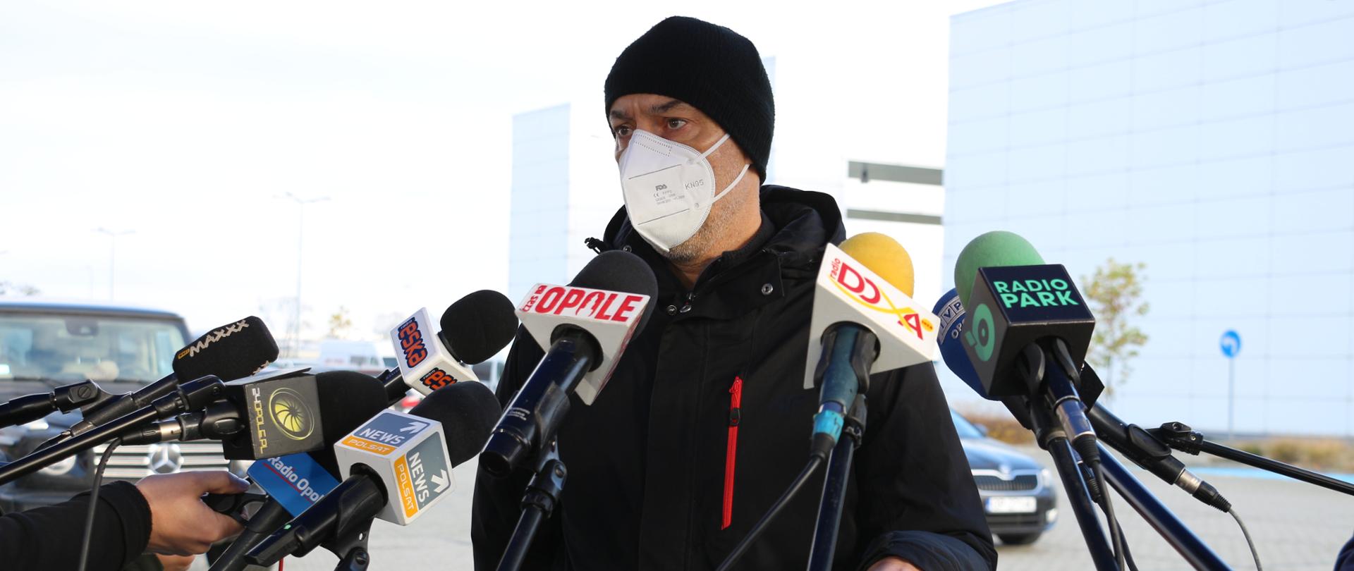 Na zdjęciu wojewoda opolski odpowiadający na pytania dziennikarzy, podczas konferencji prasowej. wojewoda znajduje się w centrum kadru. ubrany jest w czarną kurtkę, na głowie ma czarną czapkę. na przeciw niego widać kilka statywów na mikrofony dla dziennikarzy. w tle CWK w Opolu oraz stojące pod obiektem samochody osobowe. 