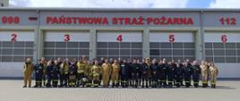 Zdjęcie grupowe po zakończonym egzaminie ze szkolenia podstawowego. Strażacy OSP ustawieni w dwóch rzędach przed garażem JRG z napisem Państwowa Straż Pożarna. Ubrani są w stroje koszarowe i specjalne koloru czarnego i piaskowego. 