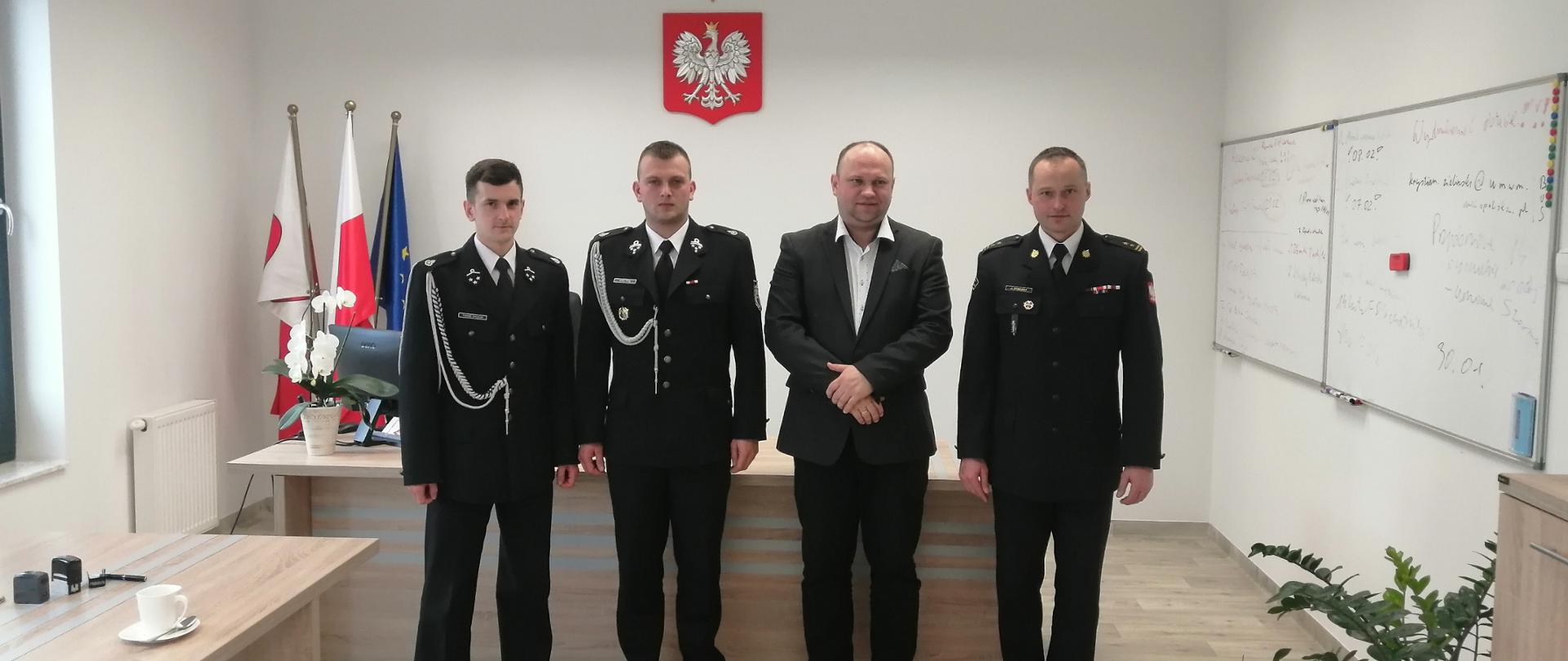 Trzech mężczyzn ubranych w czarne ubrania wyjściowe, sznury galowe, białe koszule, czarnym butami, stoą. Jeden mężczyzna ubrany w ciemna marynarkę i białą koszulę stoi. W tle na ścianie znajduje się godło polskie, oraz flagi biało-czerwone.