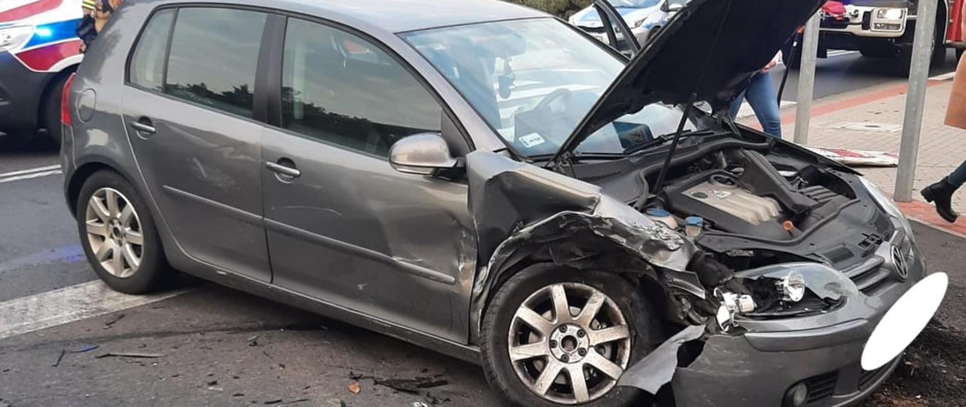 Zdjęcie przedstawia rozbity samochód marki VW Golf na tle karetki pogotowia, radiowozu policyjnego i samochodu pożarniczego.