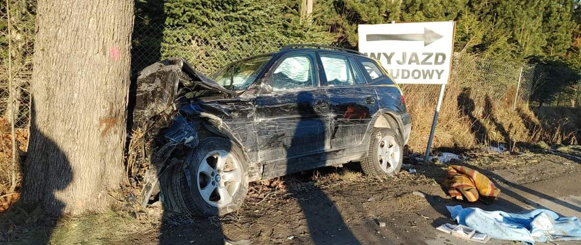 Pobocze drogi. Między tablicą informacyjną a drzewem stoi uszkodzone podczas wypadku auto osobowe. 