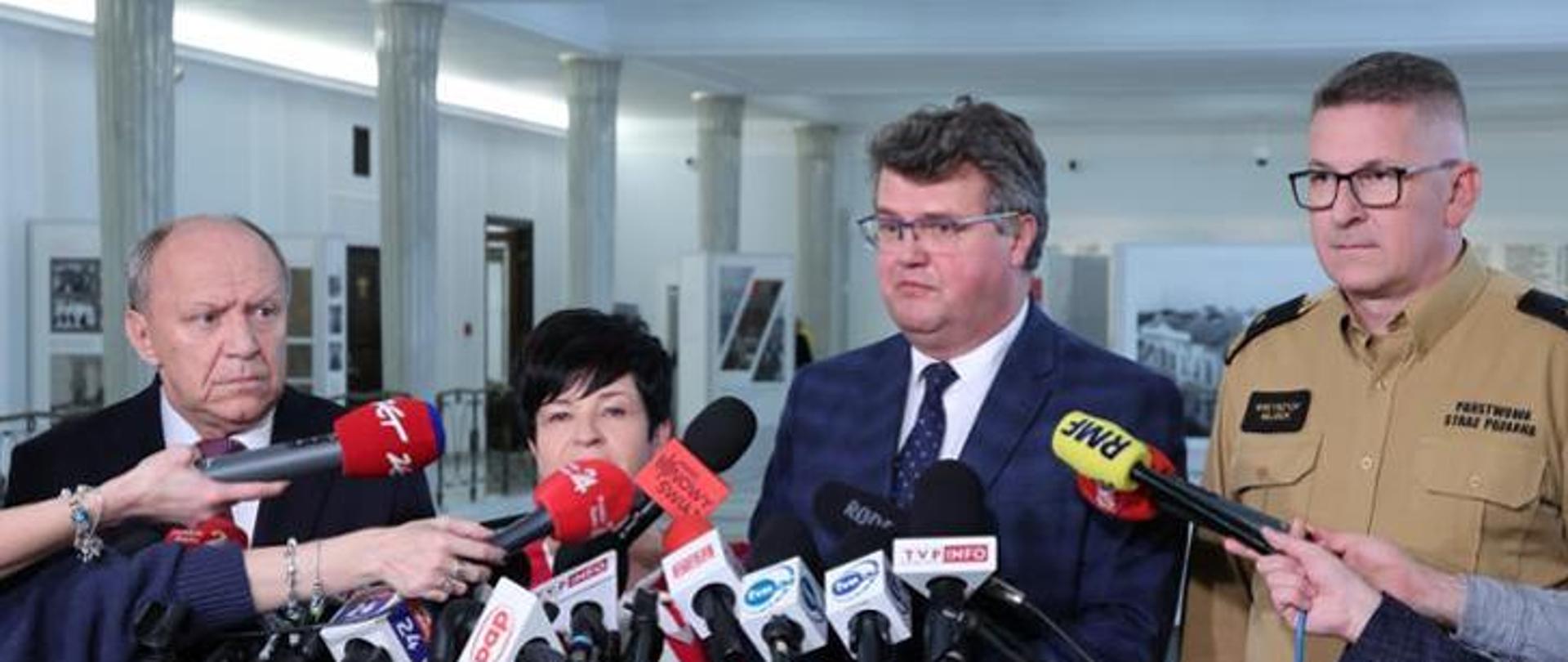 Na zdjęciu widzimy 4 osoby stojące przed mikrofonami na konferencji prasowej. 