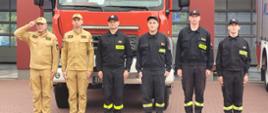 Funkcjonariusze zmiany służbowej podczas uroczystej zbiórki z okazji osiemdziesiątej piątej rocznica wybuchu Powstania Warszawskiego. Stoją w szeregu na placu komendy przed wystawionymi z garażu pojazdami pożarniczymi. Pojazdy maja włączone sygnały świetlne i dźwiękowe. Wszyscy stoją na baczność dowódca zmiany oddaje honor salutując. Dowódca zmiany i jego zastępca ubrani w służbowe umundurowanie koloru musztardowego, pozostali czterej strażacy ubrani w ubranie koszarowe koloru czarnego.