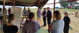 Na zdjęciu pod namiotem z drewnianych żerdzi grupa osób dzieci i młodzieży. Po prawej stronie zdjęcia stoją od lewej: kobieta w jasnej błękitnej sukience, kobieta w granatowym mundurze straży miejskiej, mężczyzna w policyjnym mundurze, kobieta w czarnym mundurze strażaka. W tle tereny zielone i elementy infrastruktury obozu harcerskiego.