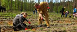 Na zdjęciu znajduje się strażak ubrany w umundurowanie służbowe, który trzyma szpadel którym kopie dół na sadzonkę, obok dziewczyna przykuca i sadzi sadzonkę drzewa. W tle widać inne osoby i dzieci, które sadzą drzewka