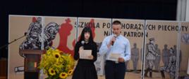 Uczennica i uczeń stoją z mikrofonami, odczytując z kartek informacje, w tle tablica z napisem: "Szkoła Podchorążych Piechoty nasz patron", po lewej bukiet żółtych kwiatów i jesienne warzywa