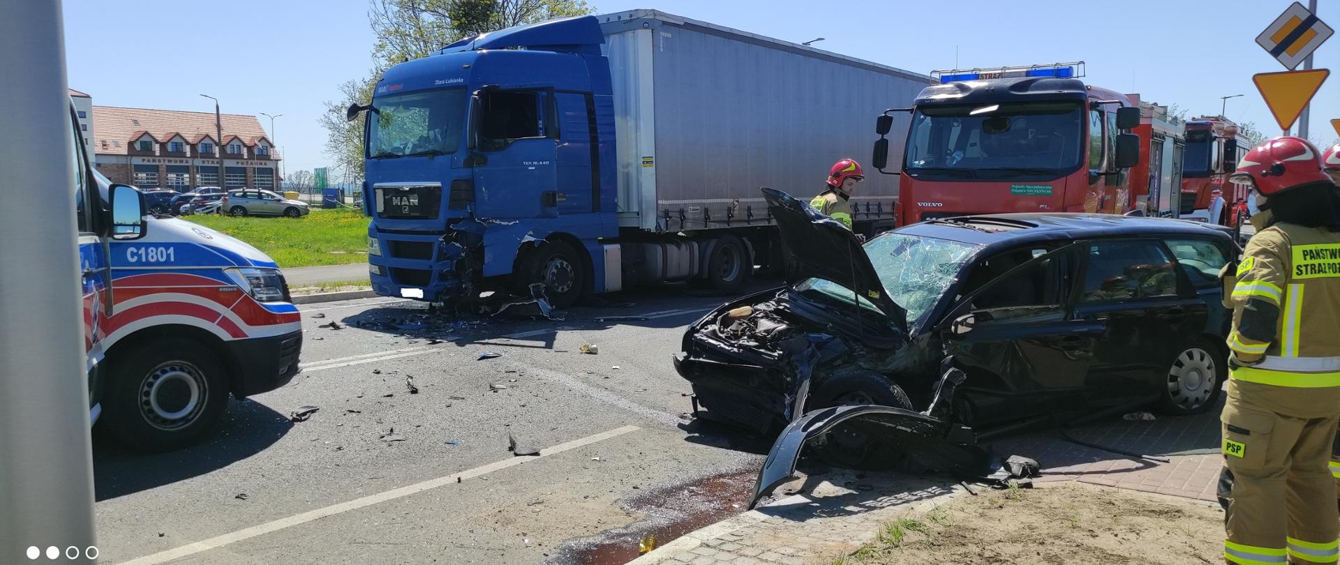 Na zdjęciu wypadek samochodu osobowego z ciężarowym. Po lewej stronie zdjęcie samochód zespołu ratownictwa medycznego. W tle samochody Komendy Powiatowej Państwowej Straży Pożarnej w Świeciu.Z prawej strony zdjęcia funkcjonariusze Komendy Powiatowej Państwowej Straży Pożarnej w Świeciu.