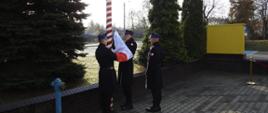 Trzech strażaków stoi przy biało czerwonym maszcie. Wciągają flagę państwową. Dookoła widać drzewa i jasne niebo. 