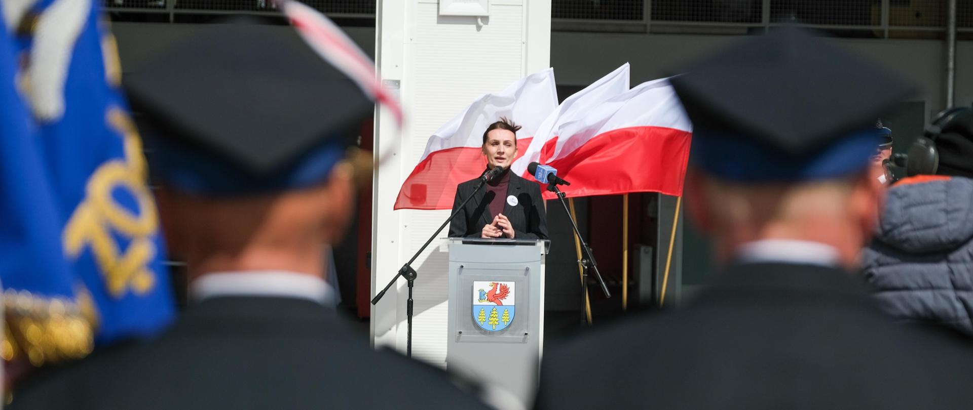 Kobieta przemawia na tle remizy OSP, przed nią stoją strażacy, w tle flagi polskie, fot. Aron Górnowicz/PSP