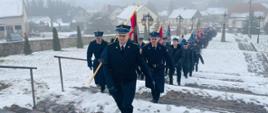 Dowódca uroczystości prowadzi poczty sztandarowe do Kościoła wchodząc po schodach. W oddali widoczne domu jednorodzinne. 