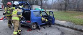 Strażacy podczas ćwiczenia z sprzętem do ratownictwa technicznego. Ćwiczą na niebieskim samochodzie fiat seicento. W tle wiata, mały murowany budynek oraz drzewa.