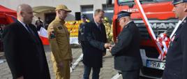 Przekazanie pojazdu ratowniczo-gaśniczego do OSP Śląskowo. Zdjęcie przedstawia plac przed remizą OSP w Śląskowie. Na tle budynku oraz uczestników uroczystości, przy przekazywanym wozie pożarniczym, burmistrz miasta i gminy Jutrosin wręcza dowód rejestracyjny pojazdu, jednemu ze strażaków OSP. Towarzyszy mu zastępca komendanta wojewódzkiego PSP w Poznaniu oraz minister.
