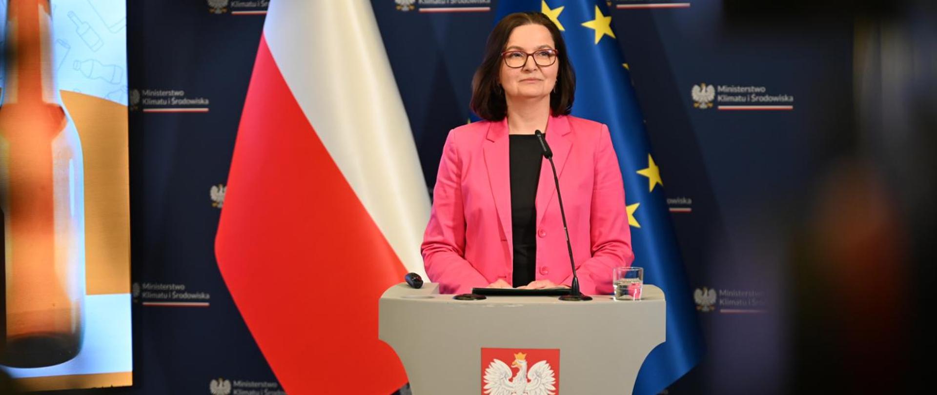 Deputy Minister for Climate and Environment Anita Sowińska during a press conference summarising the first three months of the deposit return system. In the photo, the Deputy Minister stands almost at the centre at a lectern, smiling. Behind her, on the left-hand side, part of a presentation screen featuring an image of a glass bottle is visible, and directly behind her are the flags of Poland and the European Union.
