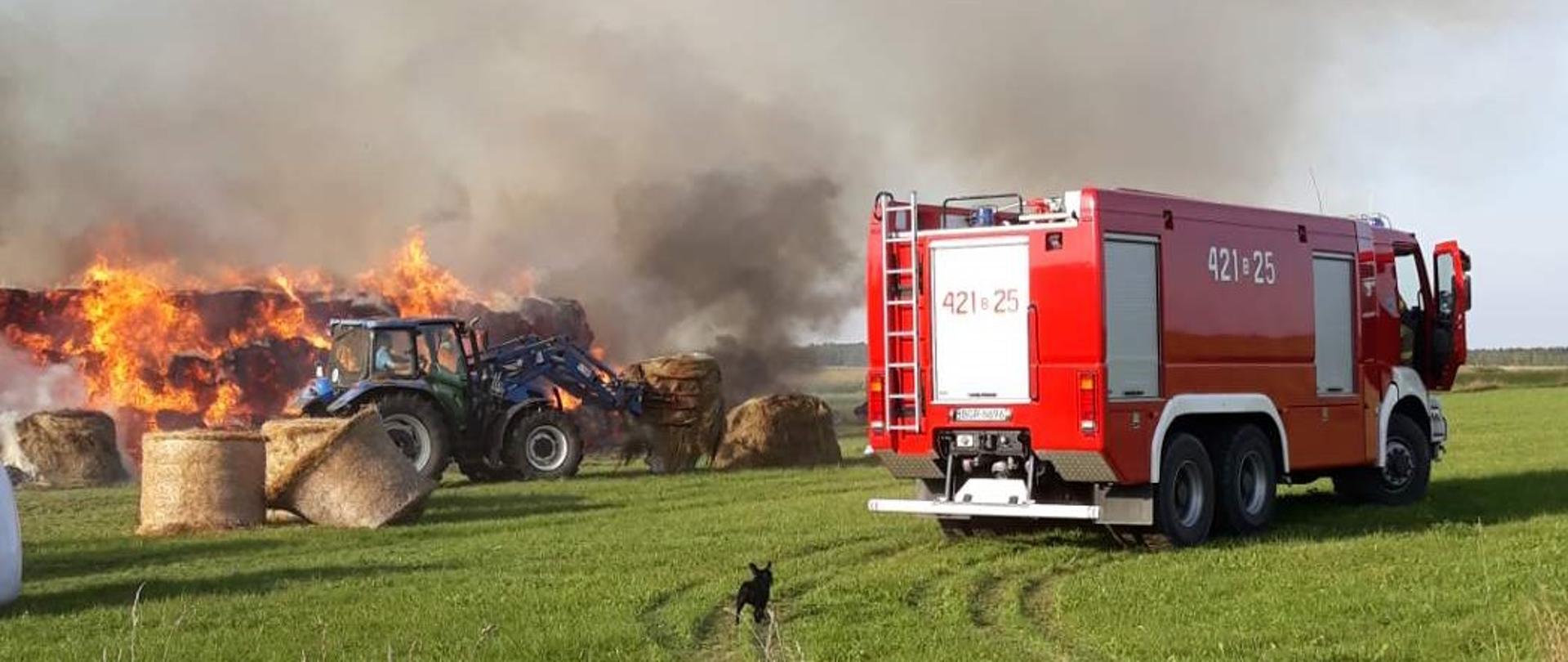 Samochód strażacki w tle ciągnik rolniczy a za nim płonąca sterta bel słomy.