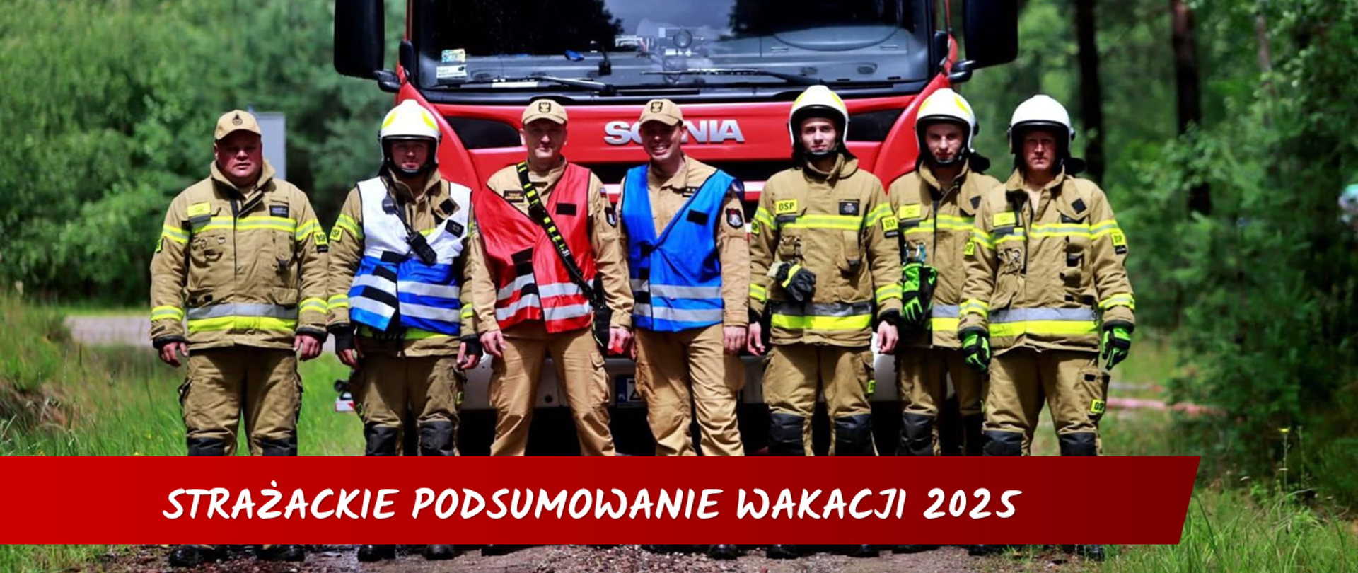 Grupa siedmiu strażaków stoi na gruntowej drodze w lesie. Za nimi widoczny jest duży, czerwony wóz strażacki z napisem SCANIA i napisem STRAŻ POŻARNA. Pojazd stoi na tle zielonego lasu.
Strażacy ustawieni są w jednym rzędzie. Wszyscy są ubrani w mundury bojowe w kolorach beżowym i khaki, z czarnymi butami. Noszą również białe i żółte kaski ochronne. Dwóch strażaków, stojących w środku, ma na sobie kamizelki odblaskowe. Jedna z kamizelek jest czerwona, a druga niebiesko-żółta. Mężczyźni stoją swobodnie, z rękami opuszczonymi wzdłuż tułowia lub skrzyżowanymi z przodu.
Scena jest jasna i dobrze oświetlona, co podkreśla szczegóły umundurowania i otoczenia. Droga jest błotnista i widać na niej kałuże, co może świadczyć o wcześniejszych opadach deszczu.
