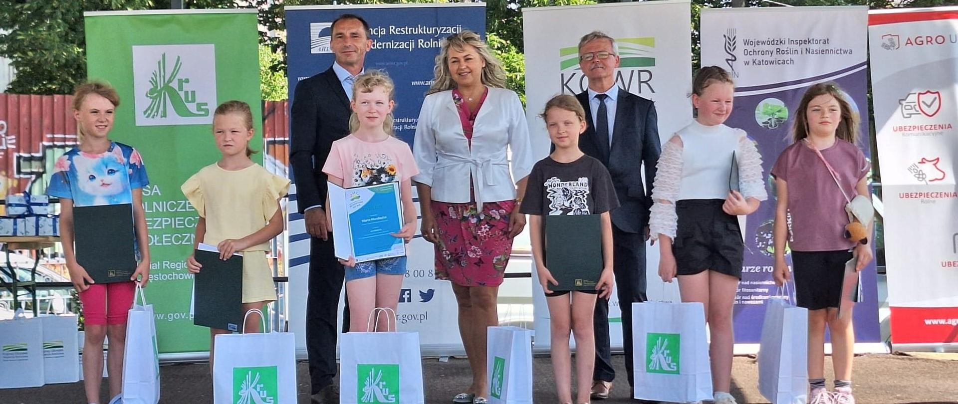 Na scenie grupa dzieci z dyplomami z tyłu stoją dwaj mężczyźni i kobieta.