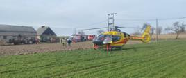 Na pierwszym planie stojący śmigłowiec LPR za nim stoją samochody pożarnicze, karetka pogotowia wokół strażacy .Przy budynkach gospodarczych stoją dwa ciągniki i maszyny rolnicze