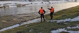 Dwóch strażaków ubranych w kapoki stoi nad brzegiem rzeki. Strażacy obserwują wzburzoną wodę. Jeden z nich wskazuje ręką na jakiś obiekt w oddali.
