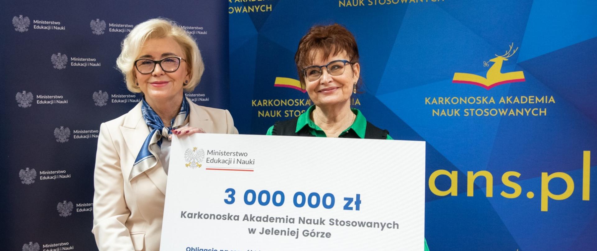 Wiceminister Machałek w białym ubraniu stoi obok ubranej na zielono kobiety trzymającej wielki symboliczny czek z napisem 3 000 000 zł, za nimi niebieska ścianka z napisem Karkonoska Akademia Nauk Stosowanych.