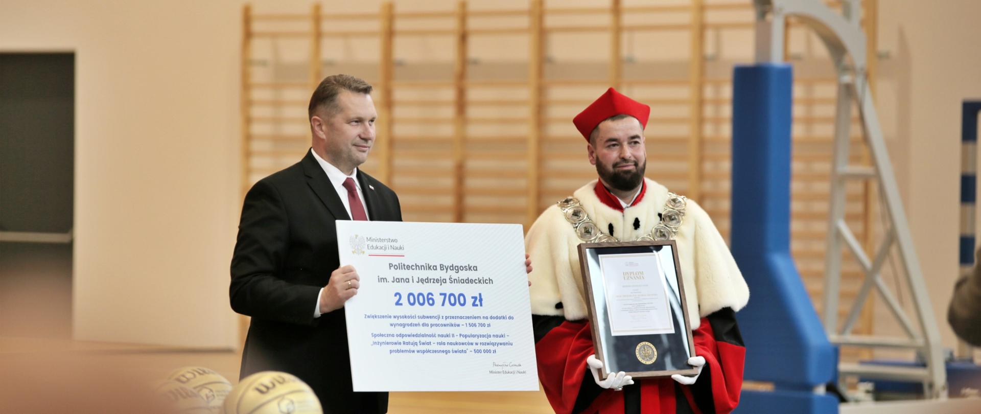 Na środku sali gimnastycznej stoi minister Czarnek, w rękach trzyma wielki symboliczny czek z napisem 2 006 700 zł, obok niego stoi mężczyzna w czerwonym ceremonialnym stroju z gronostajowym kołnierzem i trzyma oprawiony w ramki dokument.